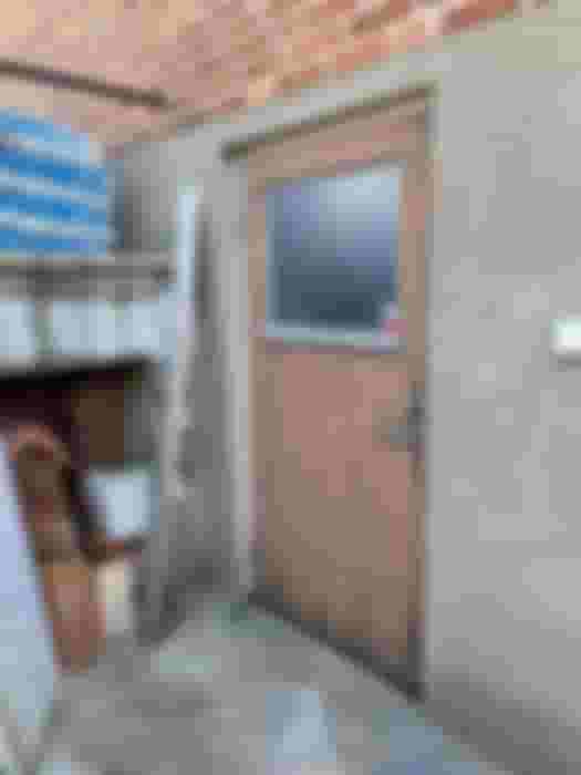 En sliten, brun dörr med fönster i en grå vägg, omgiven av byggmaterial och tegelpannor. Perfekt för renoveringsidéer.