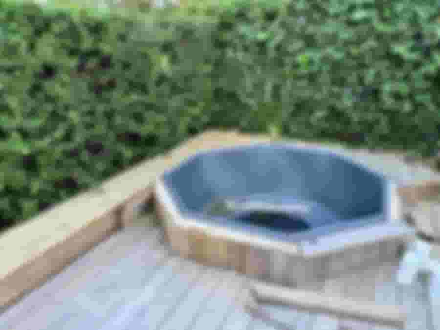 En ovala badtunna i grått material, omgiven av trädäck och grönska, med en träplanka i förgrunden. Idealisk för utomhusavkoppling.