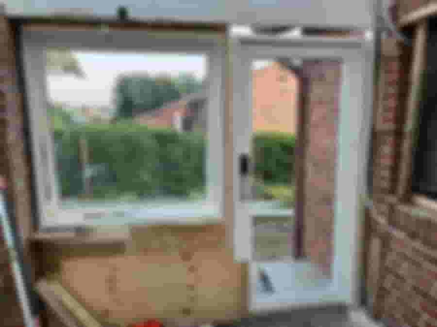 En renovering av en vägg med fönster och dörr, där utsikten visar en grönskande trädgård. Byggmaterial ligger på golvet.