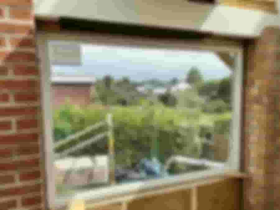 En stor, modern fönsteröppning med utsikt över en grönskande trädgård och närliggande hus, under renovering.