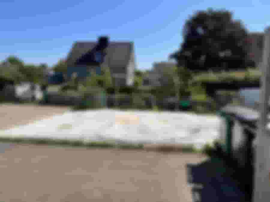 En solig tomt med en plastduk, omgiven av grönska och avfallskärl, i en lugn bostadsområde. Perfekt för framtida renoveringsprojekt.