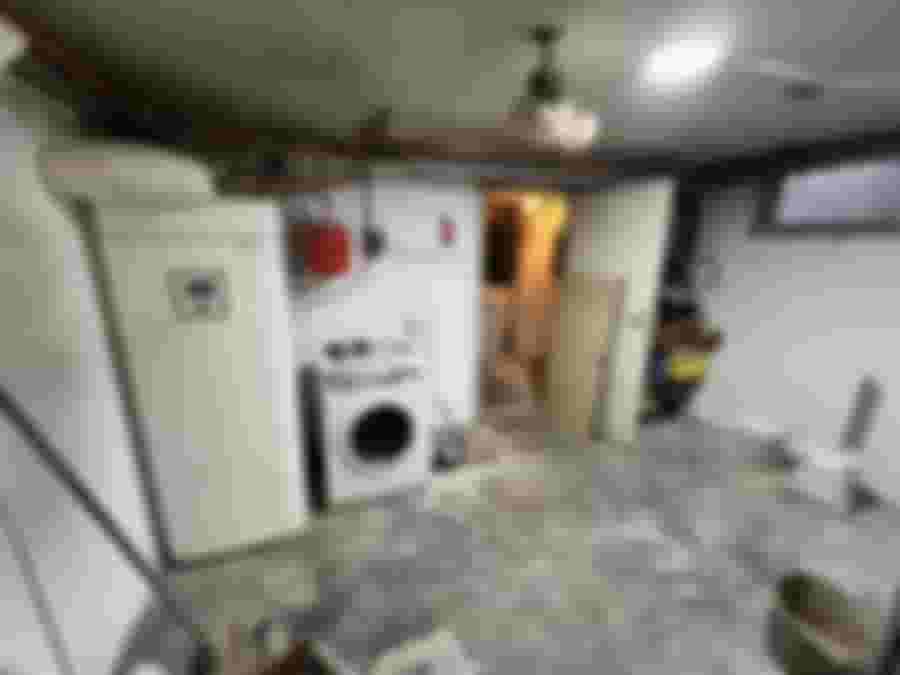 En källare med tvättmaskin och värmepump. Oinrett golv, blommor i taket, rör synliga, dörr till annat rum.