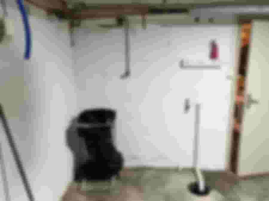 Källarrum med vit vägg, synliga rör, papperskorg och öppnad dörr. Perfekt för ett renoveringsprojekt eller inredningsförvandling.