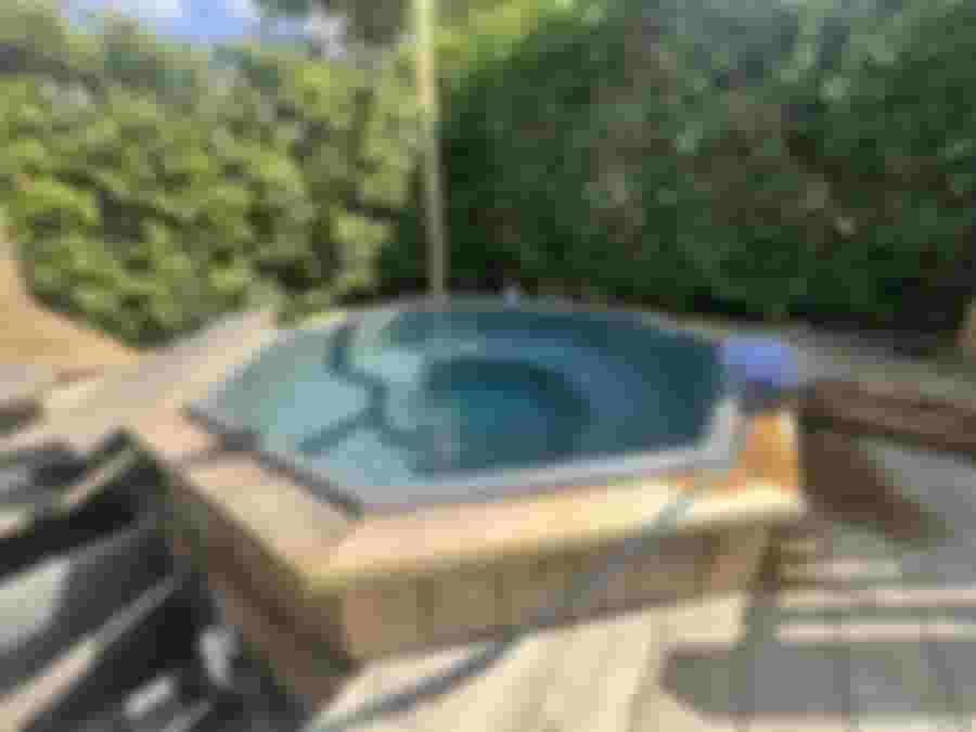 Hexagonformad jacuzzi omgiven av grönska och trädeck.
